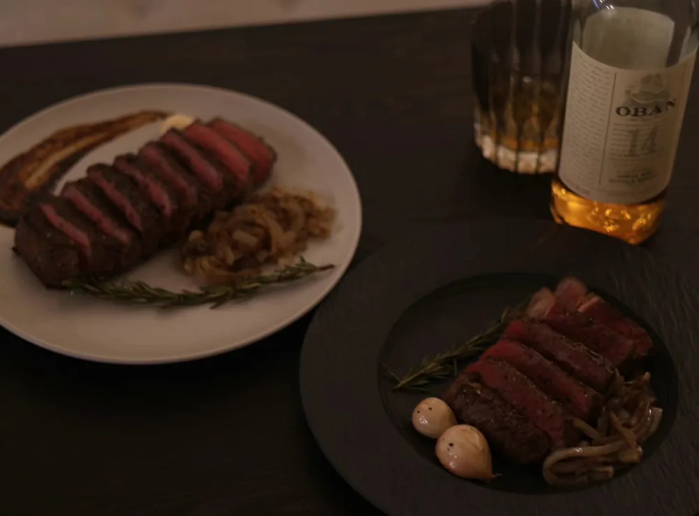 Two plates of sliced steak with caramelized onions and rosemary, beside a bottle of Scotch