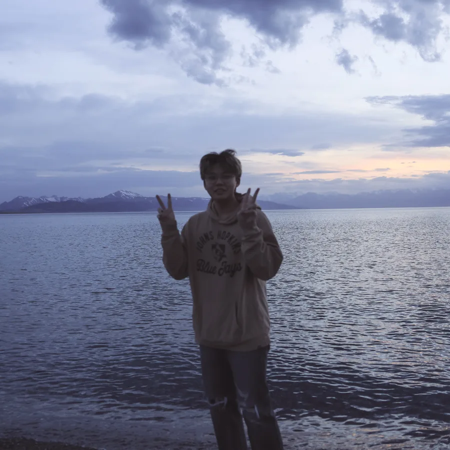 Xupeng Zhang standing by a lake at dusk with snow-capped mountains behind him